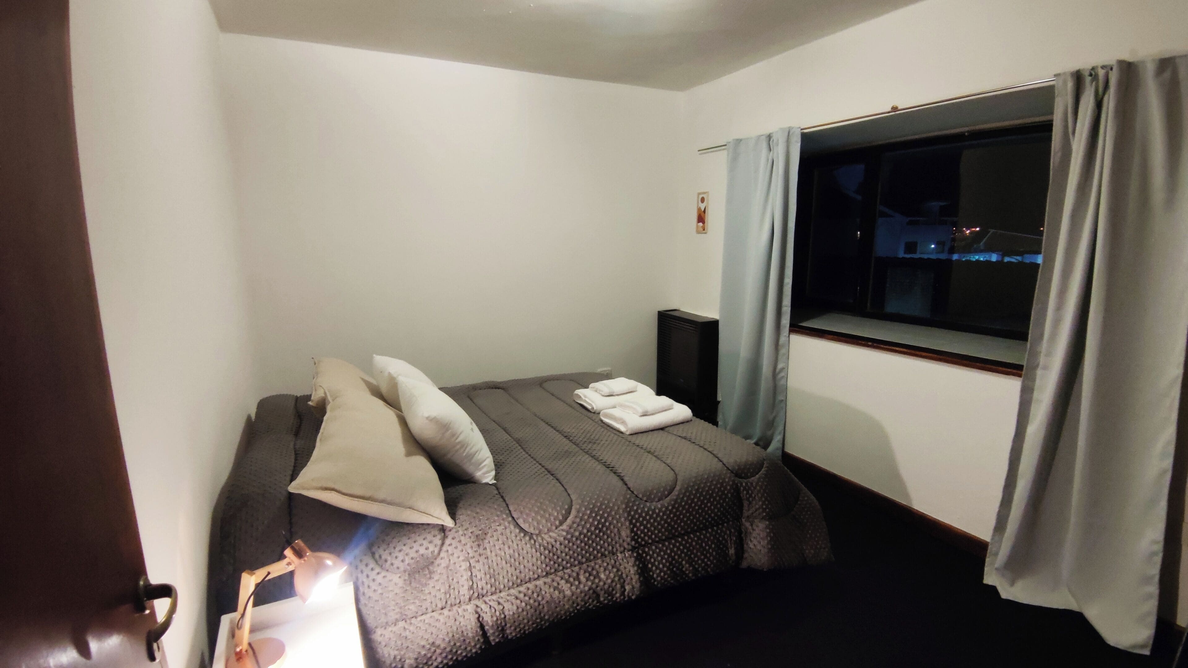 A small, dimly lit room with a single bed, a nightstand, a window with curtains, and a wall-mounted TV.