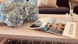Un salon moderne avec un canapé, des coussins décoratifs, un vase d’hortensias, un magazine et une petite table sur laquelle se trouve un chandelier.