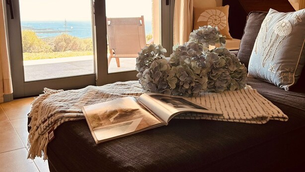 Une chambre avec une grande porte-fenêtre coulissante donnant sur un balcon, un lit avec un livre et une plante décorative, et une armoire en bois.
