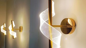 Zwei an der Wand befestigte Leuchten im modernen Design, jeweils mit einer zentralen Stange und einem dekorativen Element, das an ein Band oder einen Stoff erinnert.