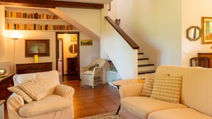 A living room with two white sofas, a wicker chair, a staircase, and a bookshelf filled with books.