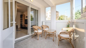 A balcony with wicker chairs and a table, a sliding glass door leading to a living area, and a view of buildings and trees. - Apartment Magic Moments (Rijeka)