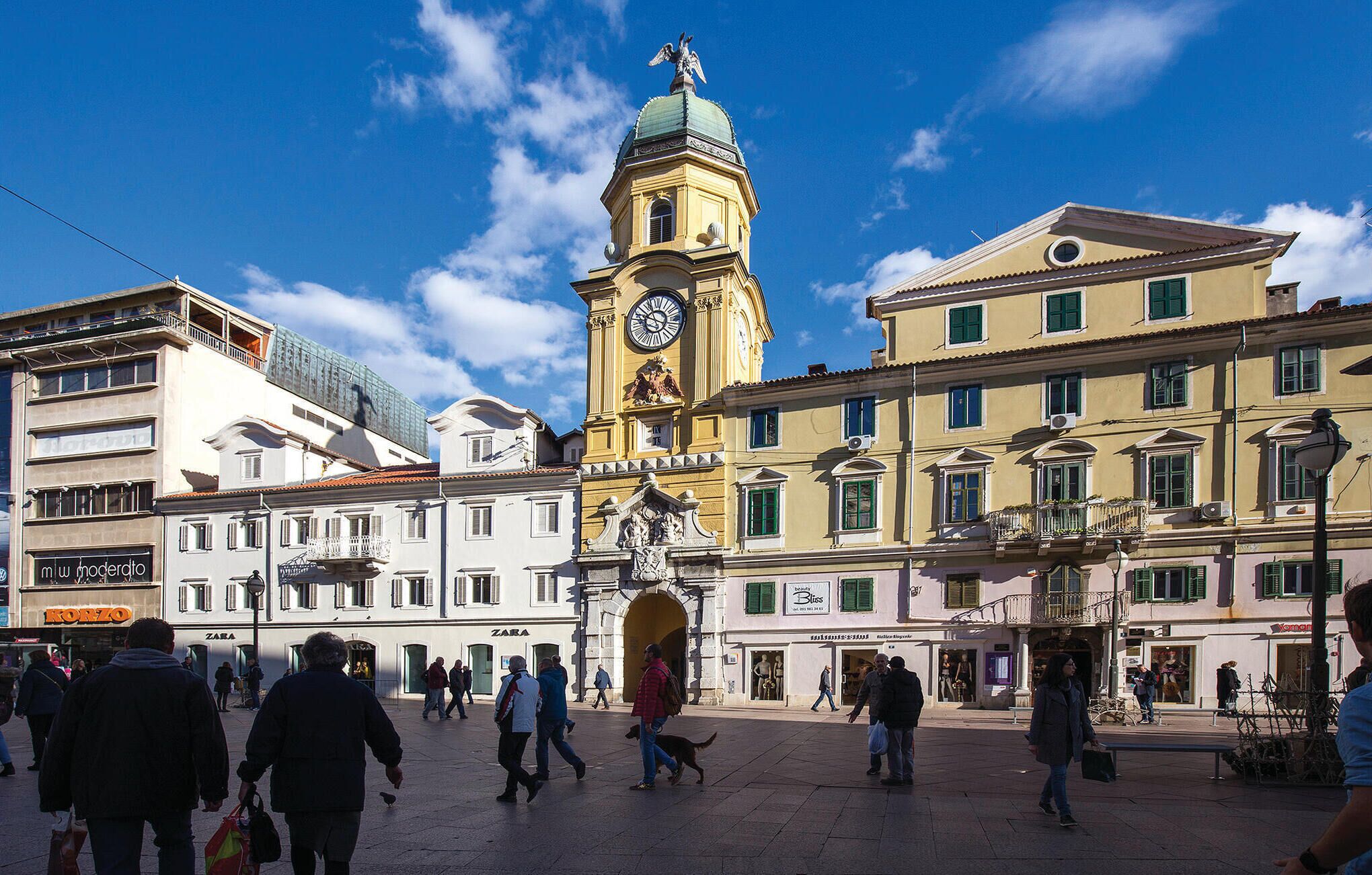 A public square with historic buildings, a prominent clock tower, and people walking.