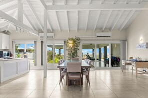 Une cuisine moderne dotée d’un grand îlot central, d’un coin repas avec une table en verre et d’une vue dégagée grâce à de grandes portes-fenêtres coulissantes.
