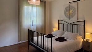 Et soveværelse med en seng, en sengetavlelampe, en fastgjort aircondition og et vindue med let gardin.