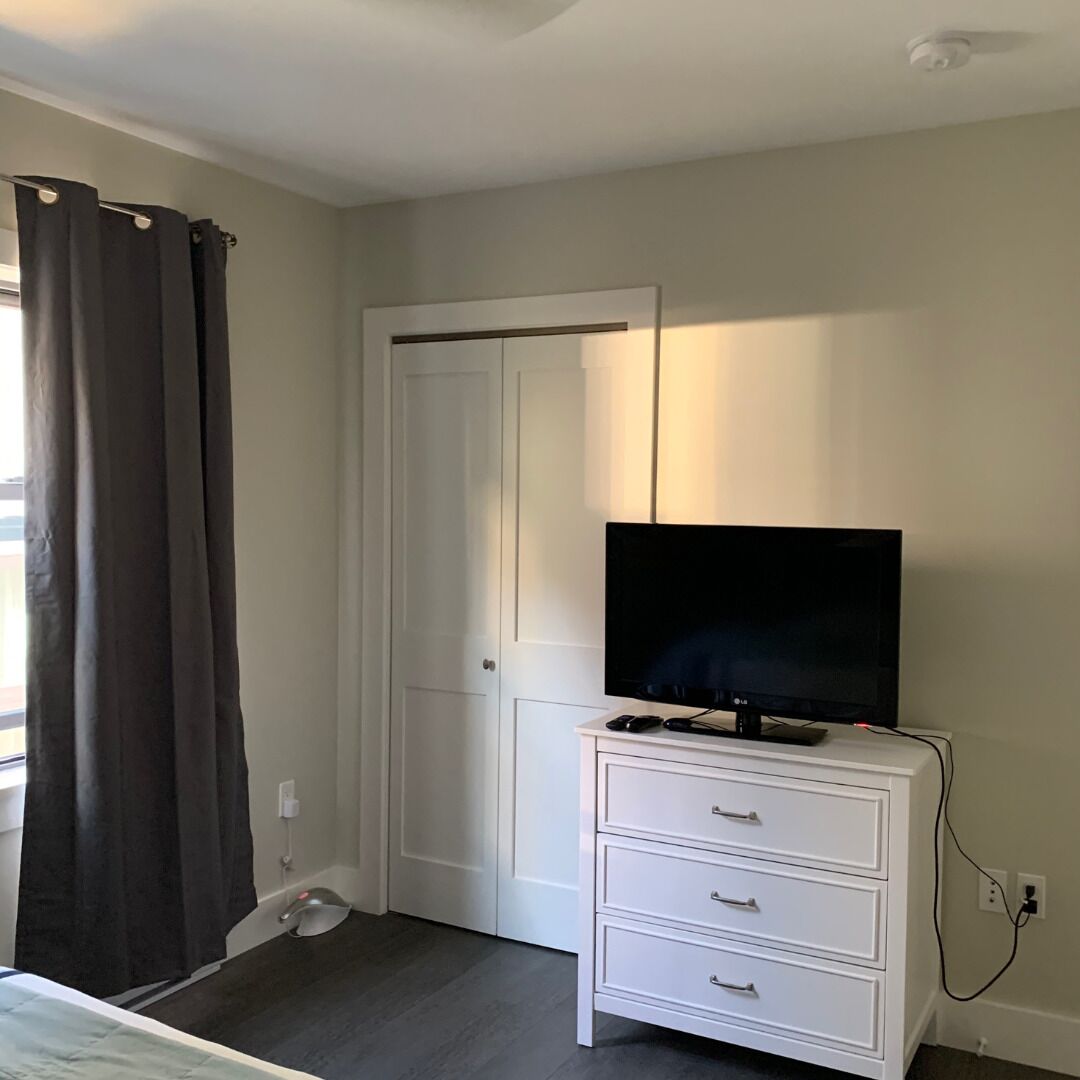 A bedroom with a white dresser, a flat-screen TV on top, a closed door, and a window with dark curtains.