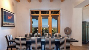 Uma sala de jantar com uma mesa comprida e cadeiras listradas, janelas amplas com vista e um quadro na parede.