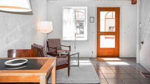A room with a wooden table, a chair, a lamp, a patterned chair, a window with curtains, and a wooden door.