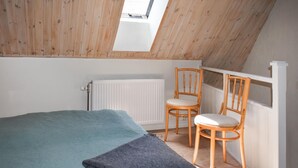 A room with a sloped ceiling, a bed with a blue blanket, two wooden chairs, and a white radiator.