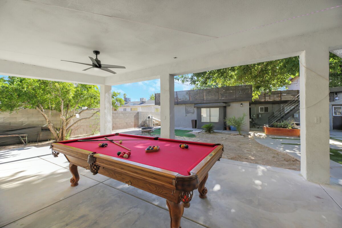 Une table de billard se trouve dans un espace extérieur abrité, doté d’un ventilateur de plafond, et est entourée d’un sol en béton. On peut admirer une maison moderne depuis cet endroit.