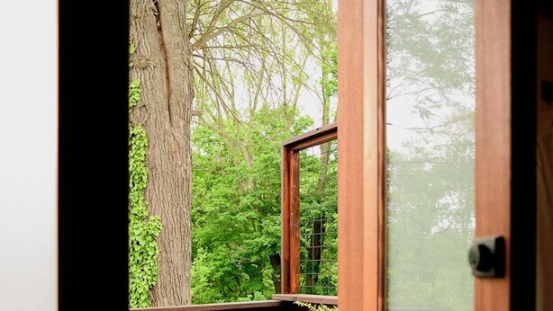 Une terrasse en bois avec deux chaises et une table, une porte vitrée et une vue sur un arbre et la verdure.