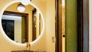 Une salle de bain avec un miroir rond, un lavabo sur colonne et une étagère en bois où sont posées des serviettes.