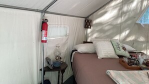 A tented room with a bed, pillows, a bedside table with a lamp, a fire extinguisher, and a window.