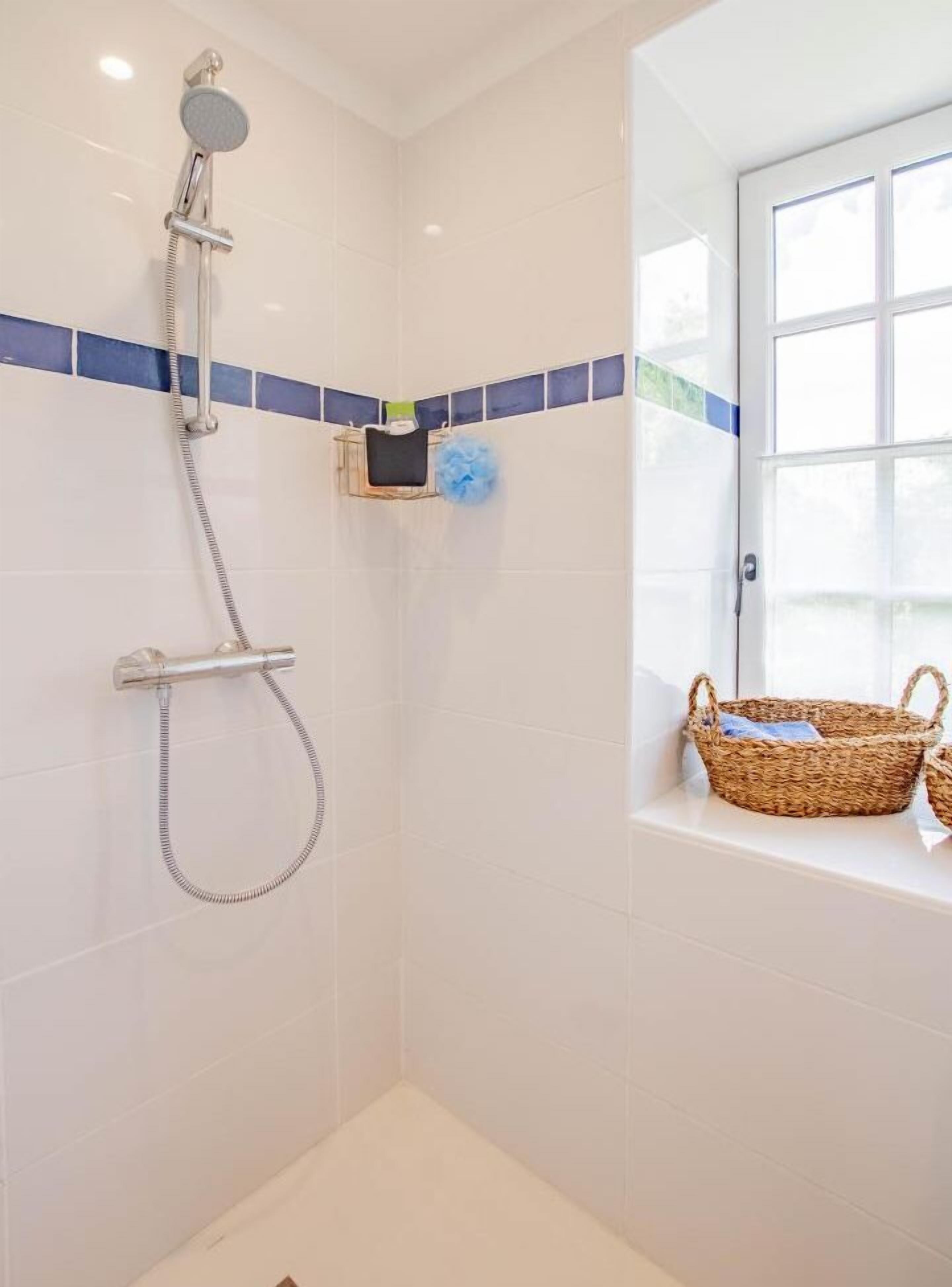 A bathroom with white tiled walls, a shower with a handheld showerhead, a small shelf with a blue bath sponge, and a wicker basket on a windowsill.