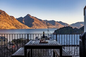 A wooden table with a bottle and glasses on a balcony overlooking a lake and mountains. - Spacious Central Executive Apartment
4 Bedroom, 2 Bathroom with amazing views (Queenstown)
