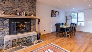 A modern living room with a stone fireplace, a dining area with a glass table, and a large window with blinds.