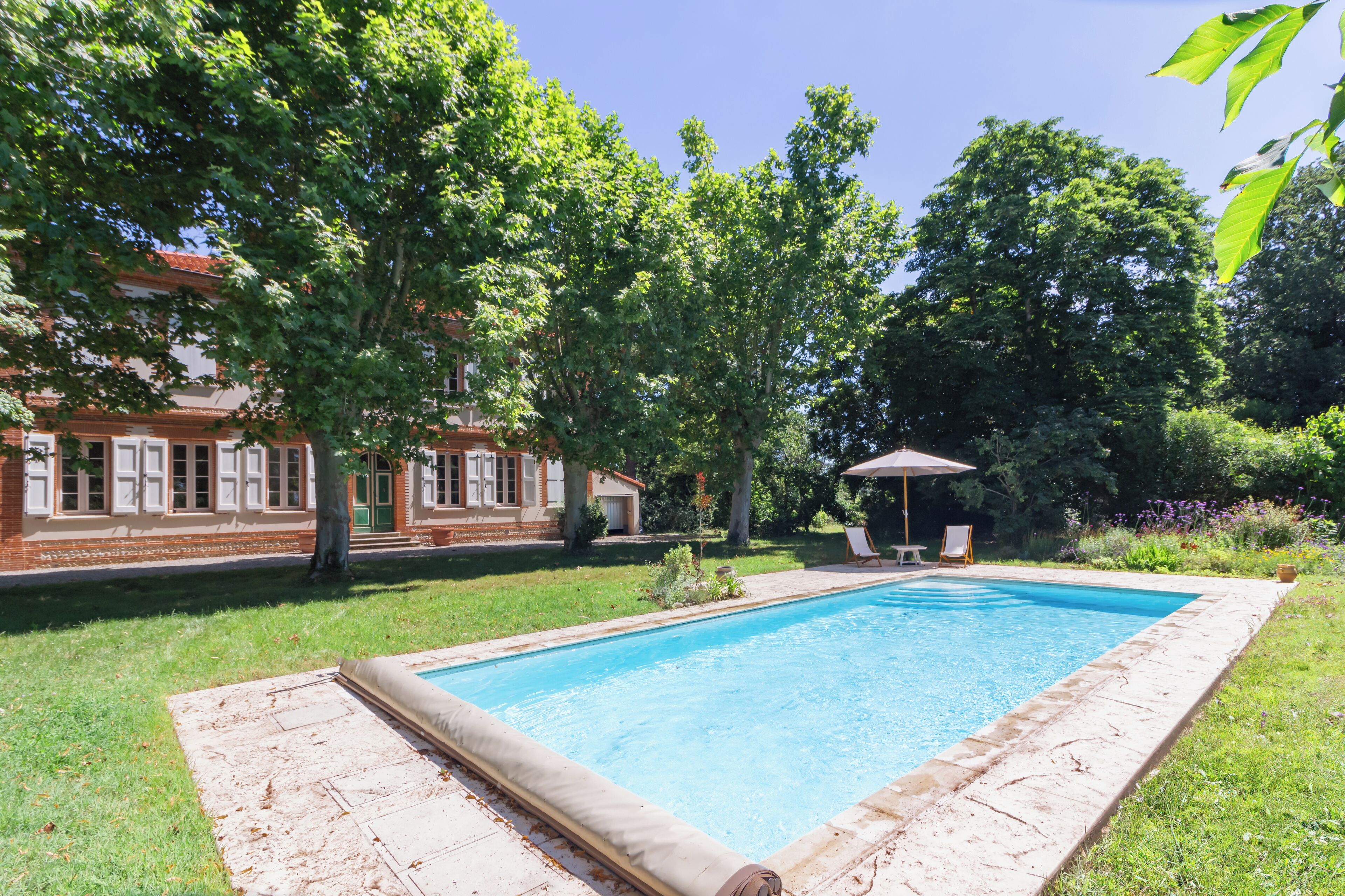 Una piscina rectangular con borde de baldosas, rodeada por un césped y árboles.