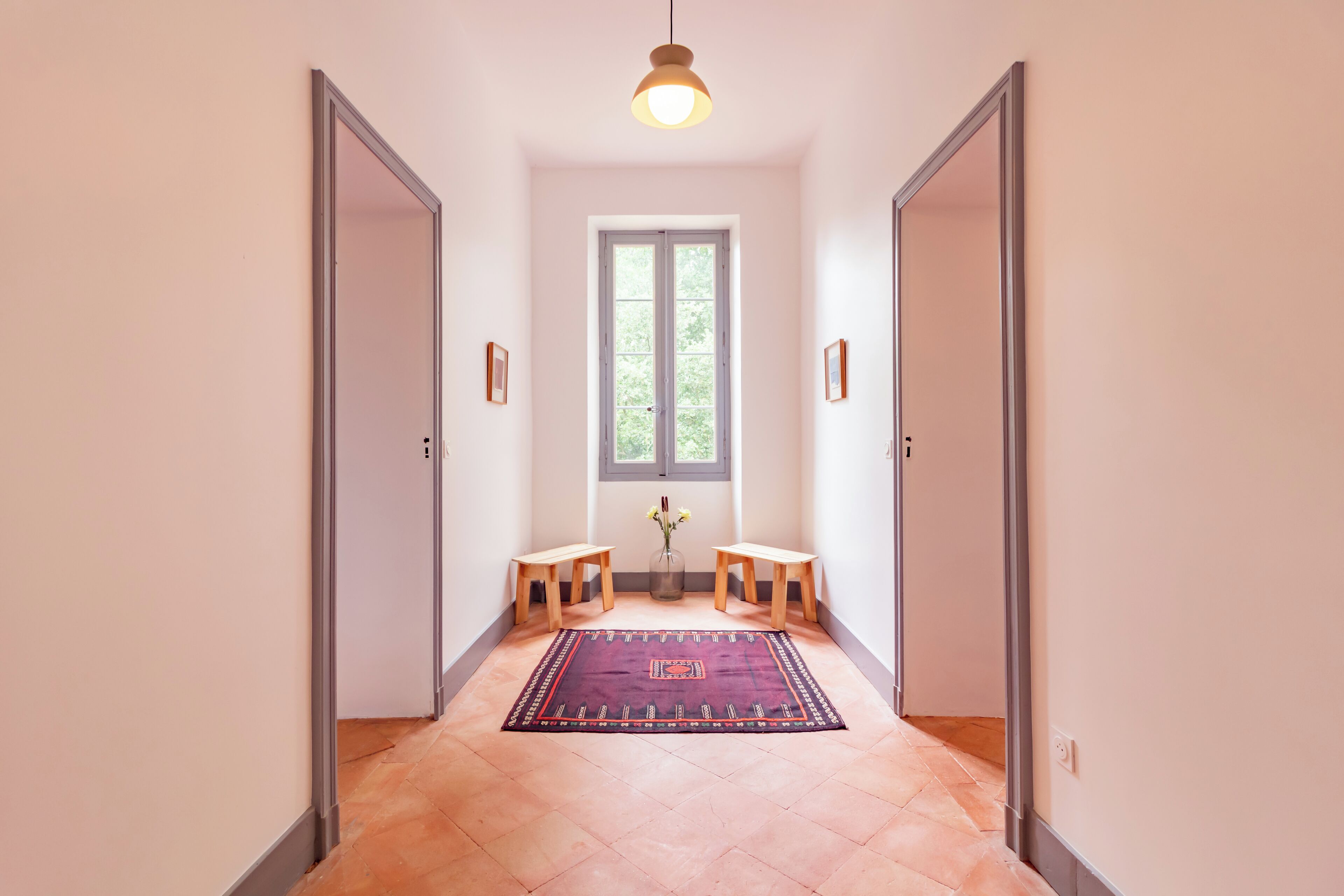A hallway with a rug, two wooden stools, a vase with flowers, and a window with a view of greenery.