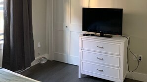 Une chambre avec une commode blanche, un téléviseur à écran plat posé dessus, une porte fermée et une fenêtre avec des rideaux foncés.