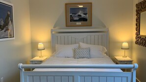 Un lit avec une tête de lit blanche et deux tables de chevet, chacune avec une lampe. Il y a un tableau encadré au-dessus du lit et un miroir sur le mur, à droite.