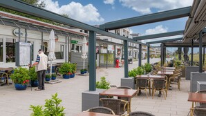 An outdoor café with tables and chairs, a person standing near a sign, and a building with a restaurant sign in the background. - 3 bedroom gorgeous apartment in Großenbrode OT Lütjenb (Großenbrode OT Lütjenb)