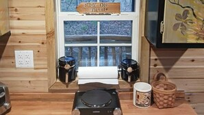A wooden cabin interior with a window displaying a 'STAY ON TRAIL' sign, a small table with a portable stove, and two black containers. - "Ridge Haus" tiny cabin on the creek at Waldbaden (Athens)