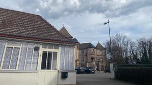 Un bâtiment avec un toit recouvert de tuiles, une grande fenêtre ornée d’un rideau à motifs et une rue en arrière-plan.