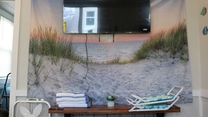 A room with a beach-themed tapestry, a flat-screen TV mounted on the wall, a wooden bench, and a chair. - Seaside Social - Steps to Wally's, Goat, L Street (Hampton)