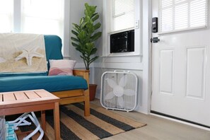 A bedroom with a bed, a bedside table, a fan, a potted plant, and a window with blinds.