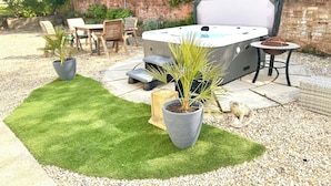 A hot tub with a built-in table and chairs, surrounded by potted plants and a patio area with a grassy patch and stone tiles. - Stunning cotswold, period country house, hot tub, great for extended family (Gloucestershire)