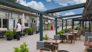 Un café al aire libre con mesas y sillas, una zona de estar cubierta y un restaurante al fondo.