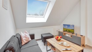 Un dormitorio tipo loft moderno con tragaluces, un sofá gris acogedor, una mesa de centro de madera con frutas y un televisor sobre un mueble de madera.