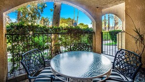 A round glass table with metal chairs, set in a covered outdoor area with a view of greenery and a clear sky. - Palm Springs 2-bedroom, 2 Bath Ground Floor condo. Garage. Pool, Hot Tub, Tennis (Palm Springs)