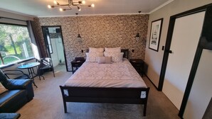 A bedroom with a stone wall, a bed with patterned bedding, a desk, a chair, and a window with a view of greenery. - Relax at Stagga Arcadia, tranquil 1 bedroom suite on rural farmlet with spa (Huiroa)
