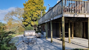 A covered outdoor seating area with a table and chairs, a wooden deck, and a large tree in the background. - Relax at Stagga Arcadia, tranquil 1 bedroom suite on rural farmlet with spa (Huiroa)
