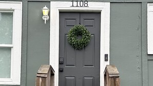 Una casa gris con una puerta gris oscuro, el número '1108' sobre la puerta, y una guirnalda colgada en la puerta.