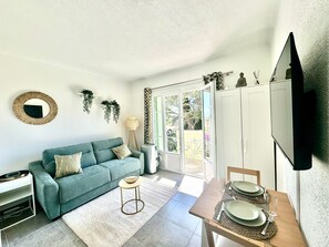 A living room with a sofa, a coffee table, a dining table set for two, and a TV mounted on the wall. - Studio Cosy à 5 min de la mer (Villeneuve-Loubet)