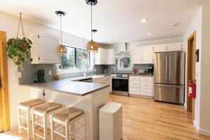 A modern kitchen with white cabinets, stainless steel appliances, a large island with bar stools, pendant lighting, and a fire extinguisher.