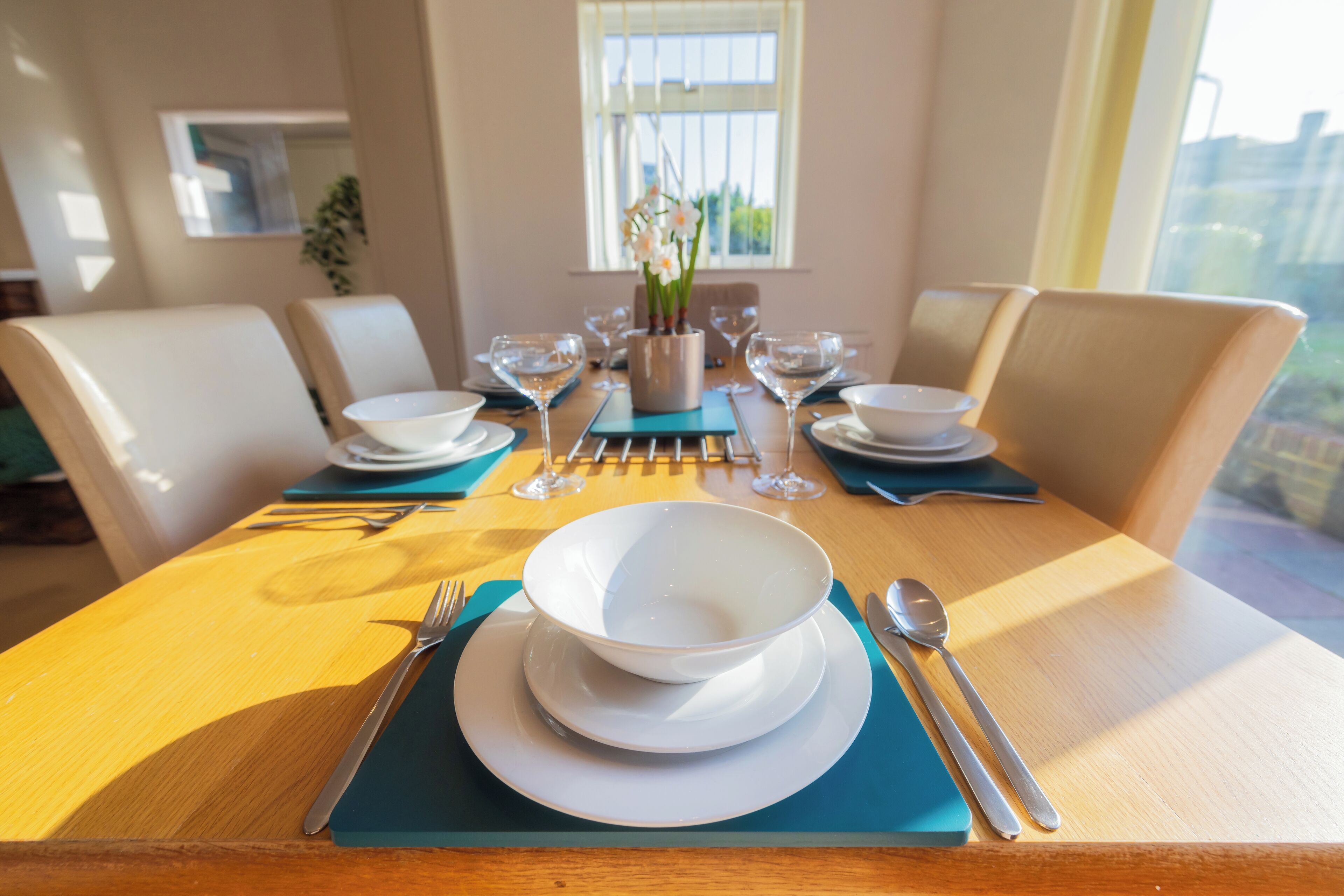 Una mesa de comedor dispuesta con platos, fuentes y cubiertos blancos, y un jarrón de flores en el centro.
