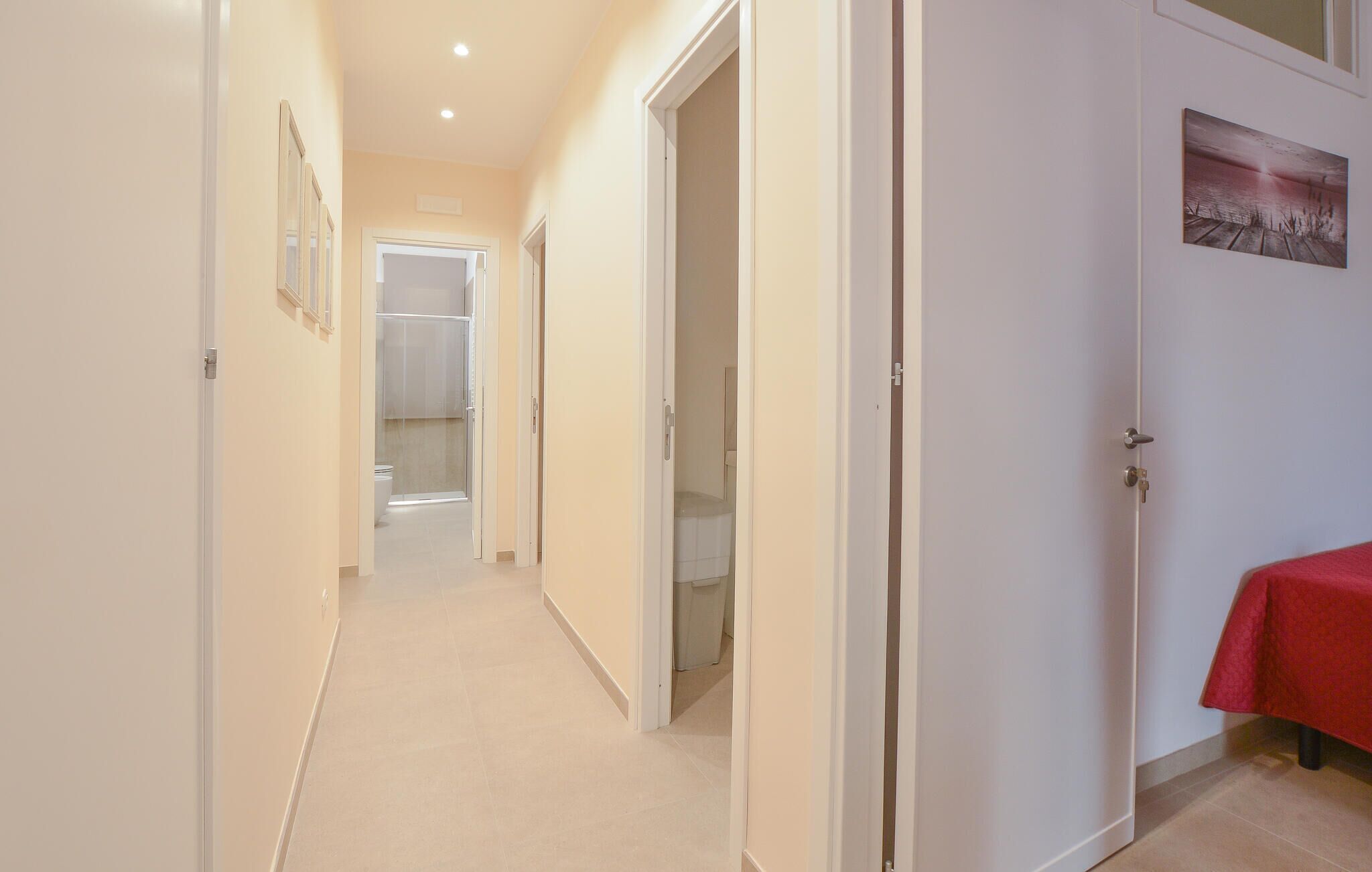 A corridor with white walls, a tiled floor, and doors leading to a bathroom and a room with a red table.
