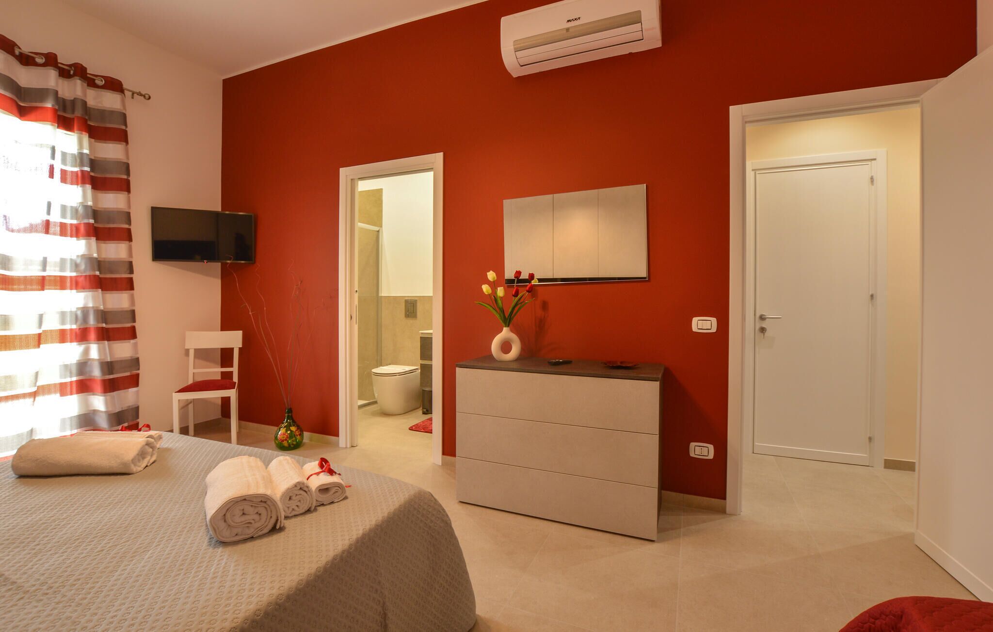 Une chambre d’hôtel avec un mur d’accent rouge, un lit avec des serviettes pliées, une commode, un téléviseur fixé au mur et une salle de bain visible à travers une porte ouverte.