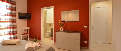 Une chambre d’hôtel avec un mur d’accent rouge, un lit avec des serviettes pliées, une commode, un téléviseur fixé au mur et une salle de bain visible à travers une porte ouverte.