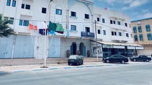 Un hôtel à la façade blanche, doté de nombreuses fenêtres et d’une enseigne indiquant qu’il s’agit d’un hôtel. Des drapeaux sont accrochés à un mât devant l’immeuble.
