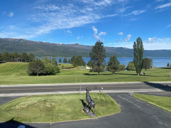 A grassy area with a deer sculpture, a road, and a lake with mountains in the background.