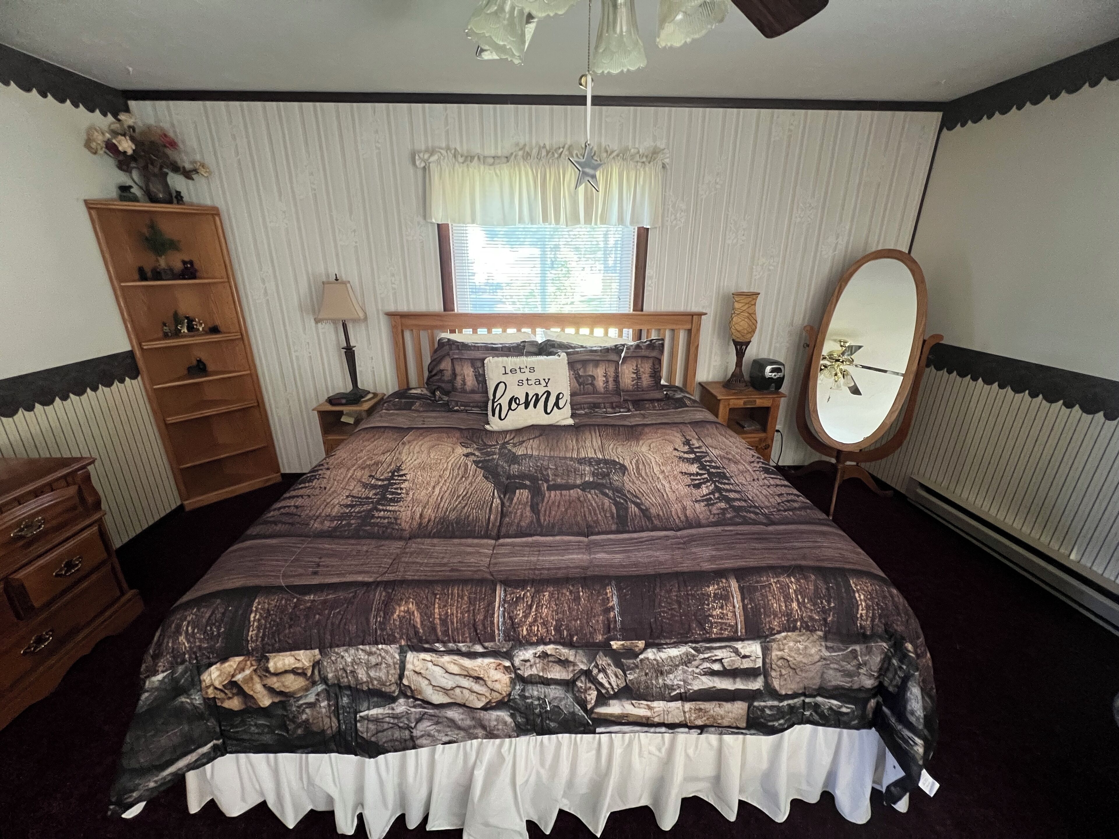 Une chambre à coucher avec un grand lit, une table de chevet en bois, une commode, un miroir, une fenêtre avec des rideaux et une étagère fixée au mur.
