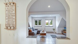 Ein modernes Wohnzimmer mit einer Couch, einem Couchtisch und einem Fernseher. Zwei Fenster sorgen für natürliches Licht und bieten einen Blick auf Grünflächen.