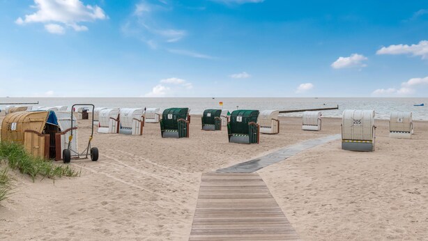 Ein Strand mit mehreren Reihen von Liegestühlen und Sonnenschirmen, einer Holzpromenade und dem Meer im Hintergrund.