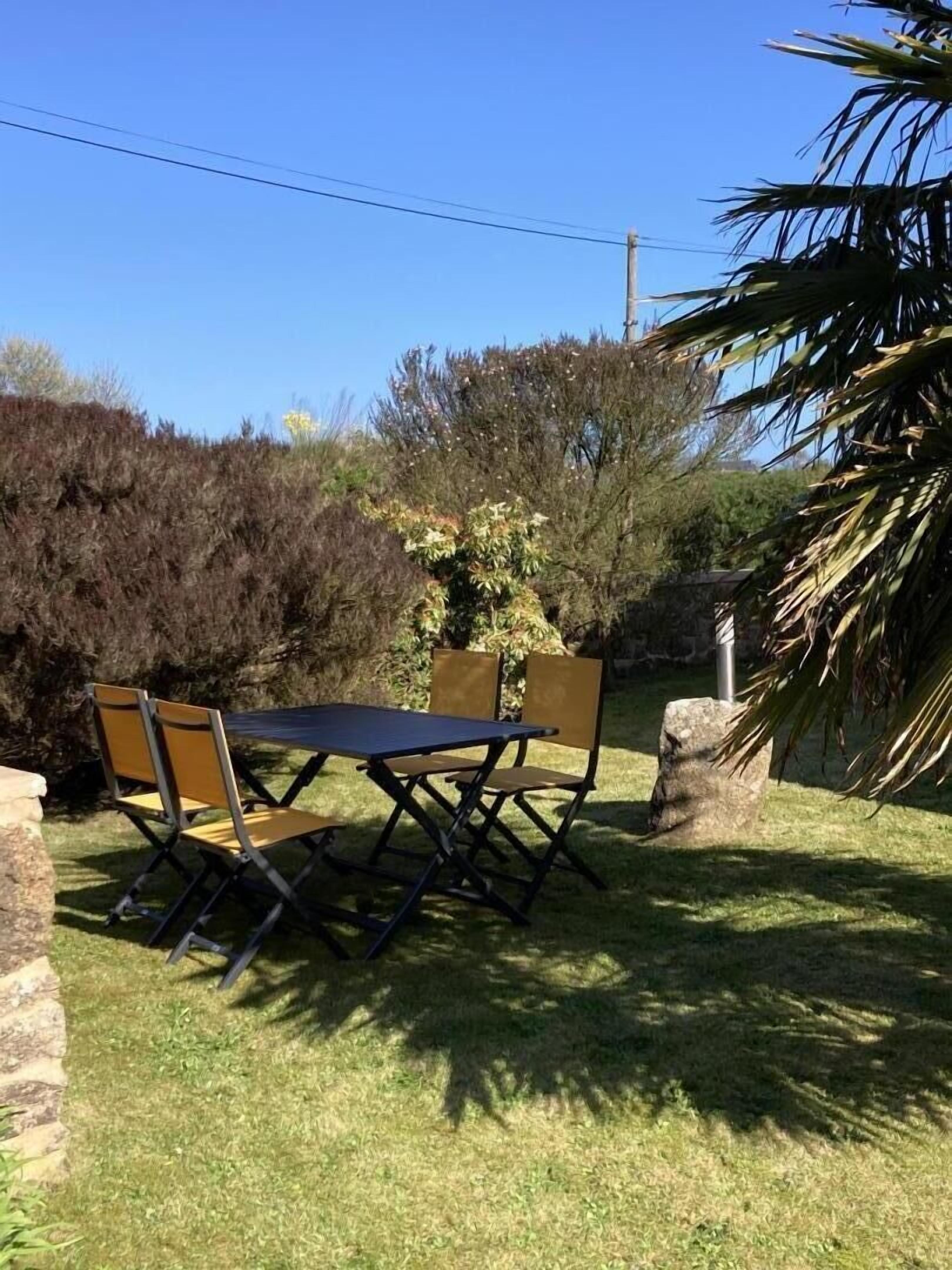 Een terras buiten met een tafel en stoelen, omgeven door gras en diverse struiken.