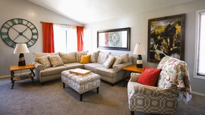 A living room with two sofas, a coffee table, and a patterned armchair. There are two side tables with lamps, a large wall clock, and a framed picture.