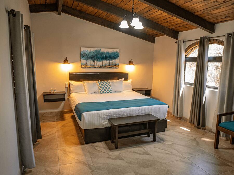 A bedroom with a bed, bedside tables, a bench, a chair, a painting, and a window with curtains.